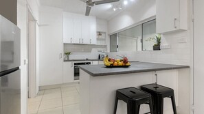 Une cuisine moderne avec des meubles blancs, un îlot central doté de tabourets, un four encastré et un panier à fruits.
