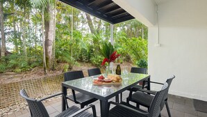An outdoor patio with a glass table and black chairs, surrounded by lush greenery and a small water feature. - 99 Reef Resort  |  Port Douglas (PORT DOUGLAS)