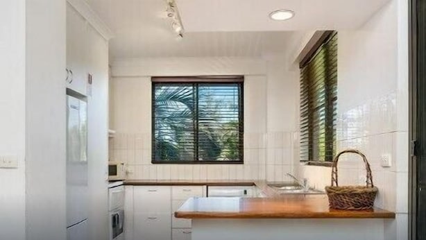 A modern kitchen with white cabinets, a wooden countertop, a window with blinds, and three stools. - Breeze at Byron 1 Clarkes Beach (Byron Shire)