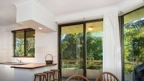 A modern kitchen with a central island, wooden stools, and a large sliding glass door leading to a balcony with a view of greenery. - Breeze at Byron 1 Clarkes Beach (Byron Shire)