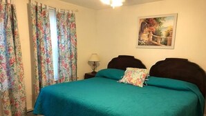 Ein Schlafzimmer mit einem Bett, floralen Vorhängen, einem Nachttisch, einer Lampe und einem Bild an der Wand.