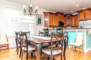 Uma sala de jantar com mesa e cadeiras de madeira, um lustre, armários de madeira e uma cozinha com eletrodomésticos.