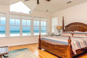 Quarto com uma grande janela com vista para a praia, uma cama de madeira e uma mesa de cabeceira com abajur.