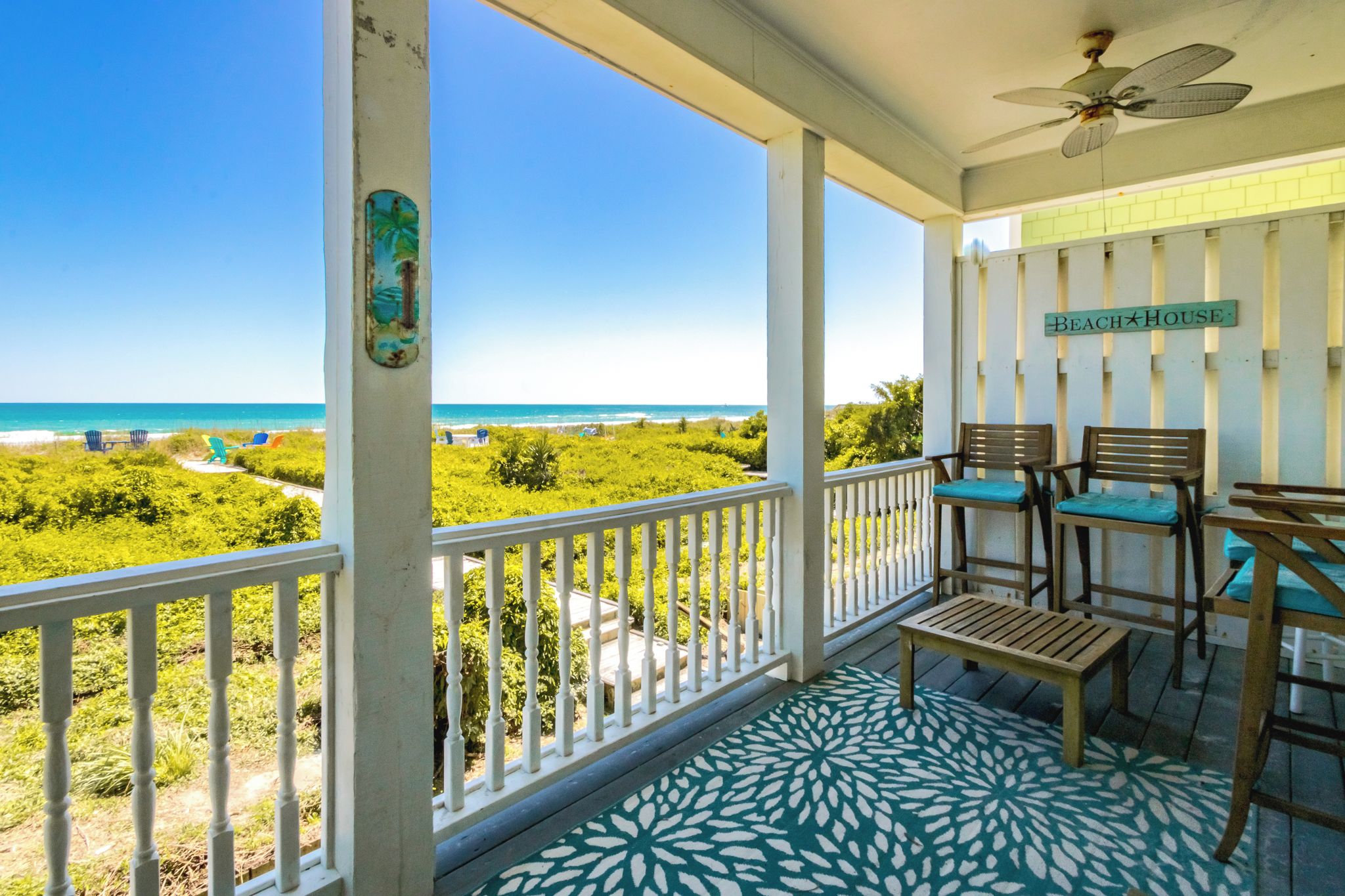 A balcony with a view of the beach, a sign reading 'Beach House', and outdoor furniture.