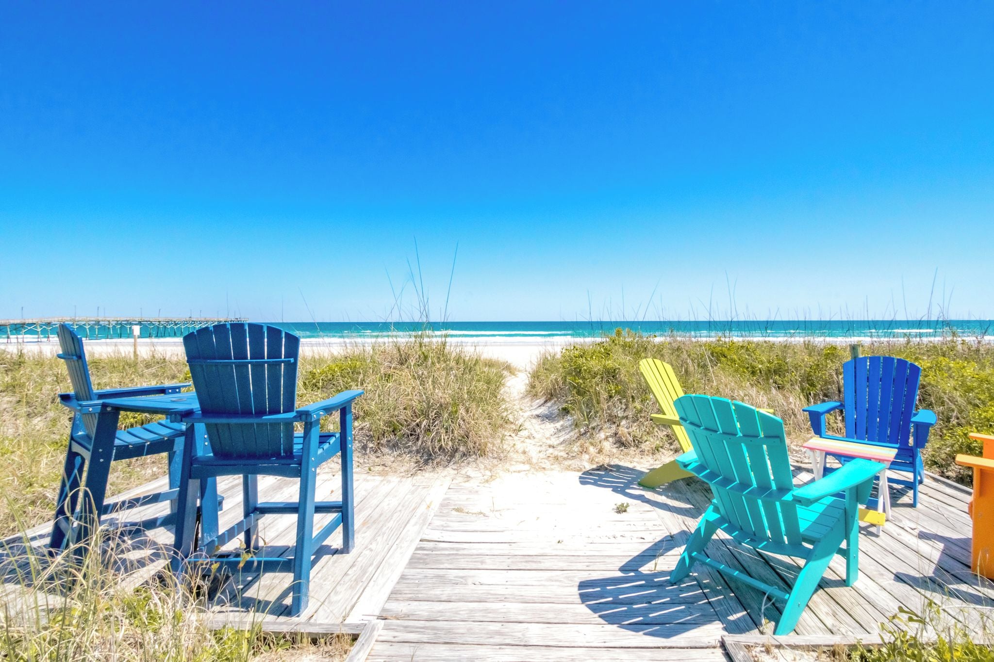 A wooden boardwalk leading to a beach with several colorful Adirondack chairs.