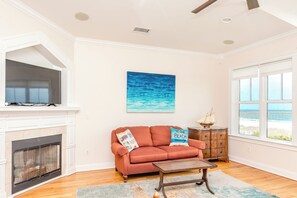 A living room with a fireplace, a sofa with beach-themed pillows, a painting of the ocean, and a view of the beach through a window. - Beach Therapy - Oceanfront!! 4 BR / 3 BA Enjoy the views from Master Bedroom! (Topsail Beach)