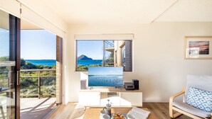 A modern living room with a large window overlooking the sea, a flat-screen TV, a coffee table with books and a coffee pot, and a comfortable chair. - Intrepid, 15 3 Intrepid Close - right on the beachfront and WiFi (Nelson Bay)