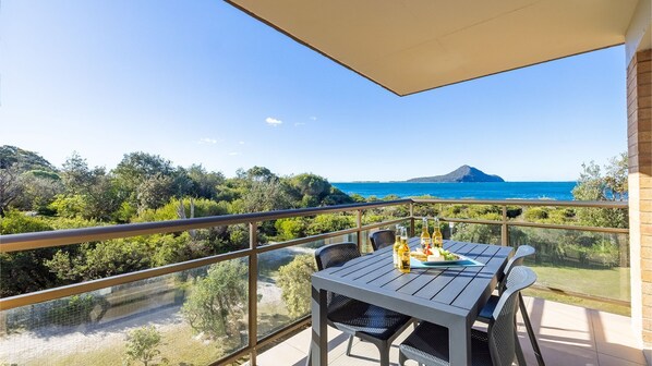 Un balcón con una mesa dispuesta para dos personas, con vista a un paisaje costero con árboles y una montaña a lo lejos.