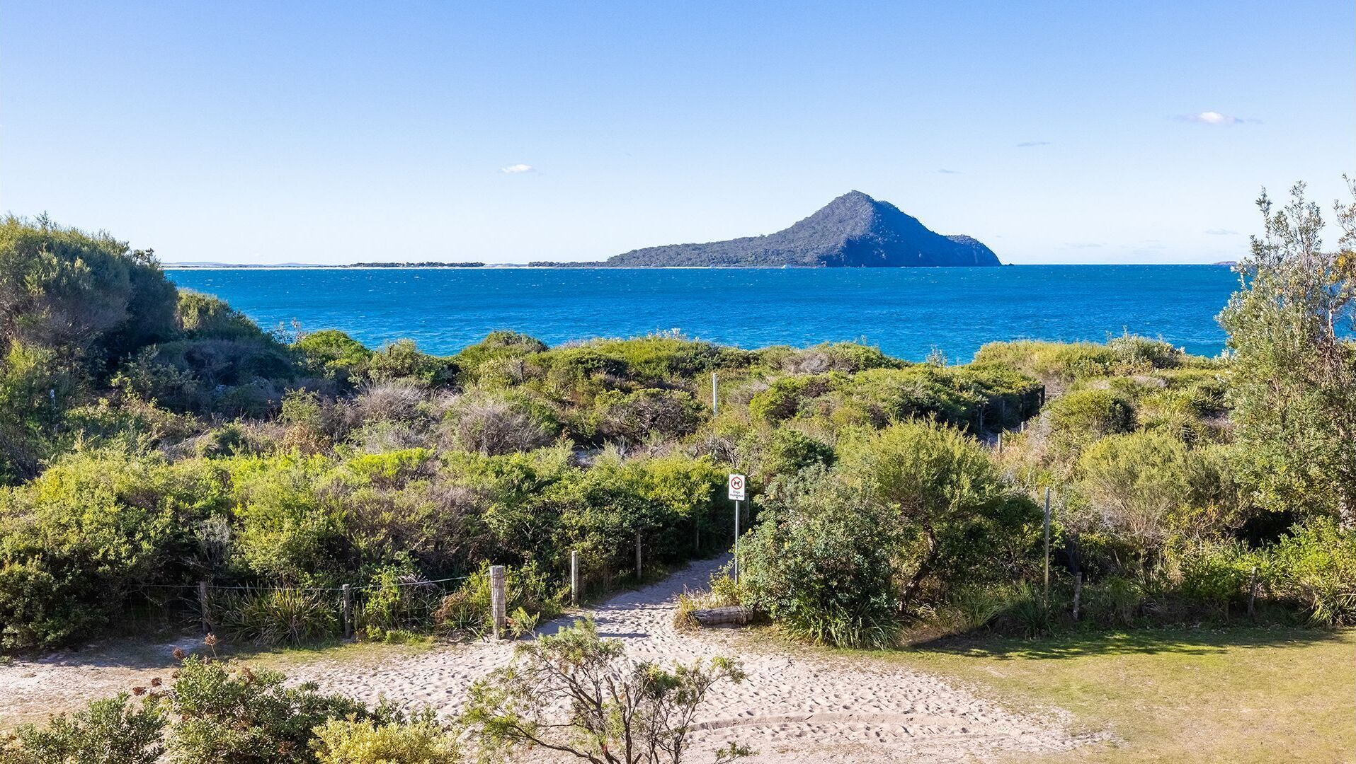 A coastal path leading to a beach with dense vegetation and a clear view of the sea and a distant island.
