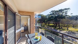 Un balcón con una mesa para dos, una puerta de vidrio que da a un dormitorio y vistas a un parque con árboles.