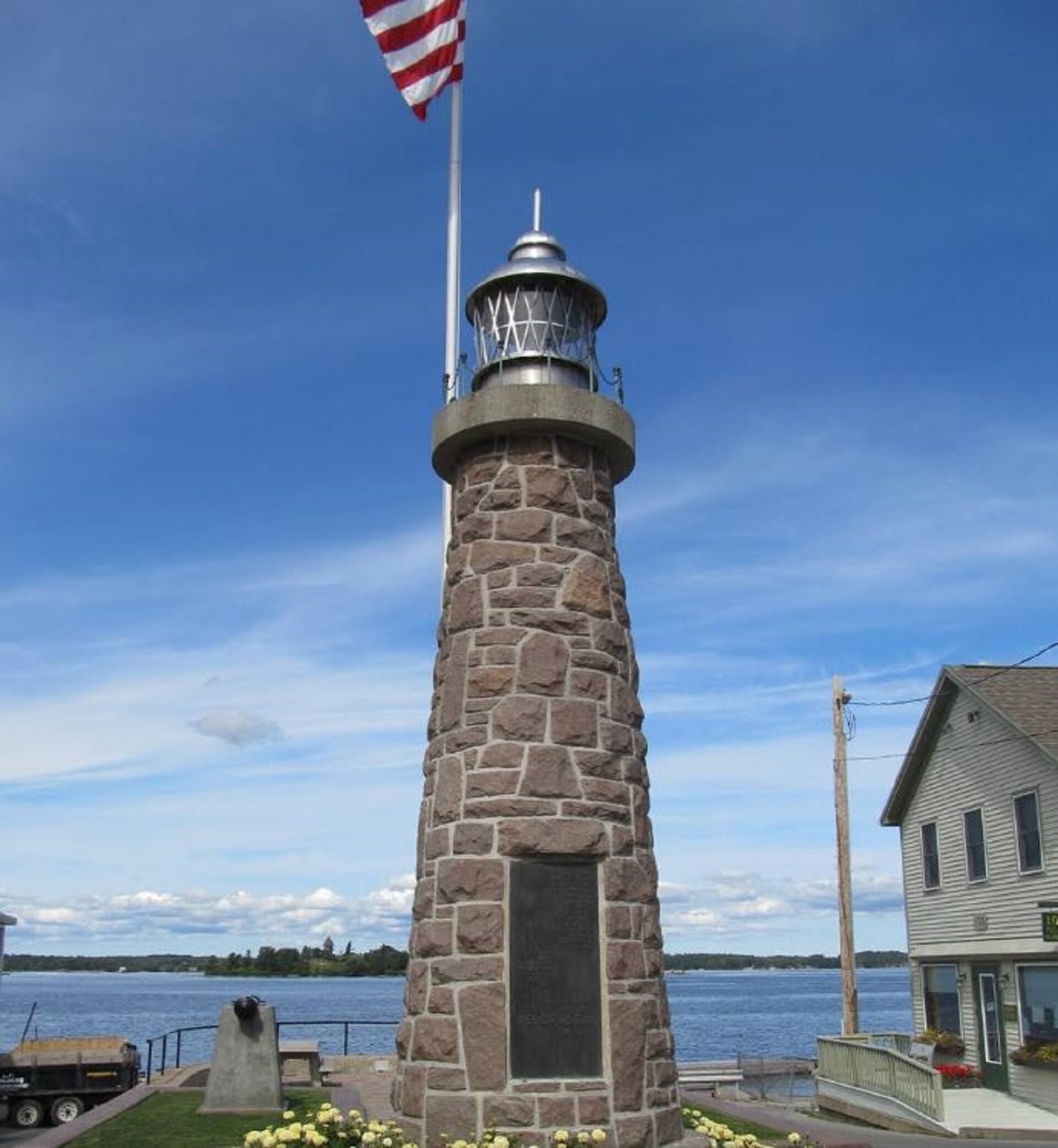 Un faro con base in pietra e sommità in metallo, con una bandiera americana in cima, e un corpo d'acqua sullo sfondo.