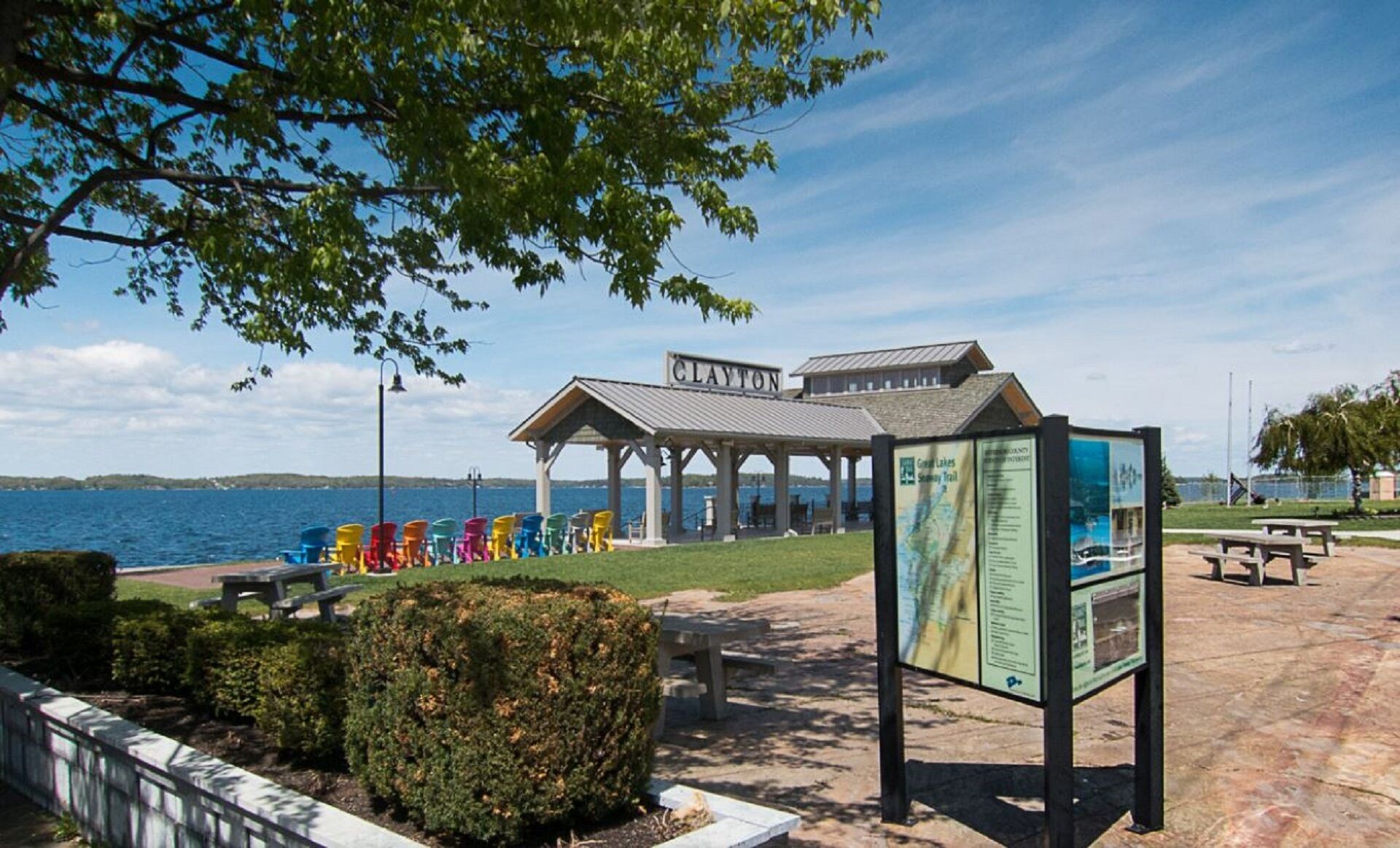 A pavilion with a sign reading "Clayton" by a lake, colorful chairs, and a picnic area.