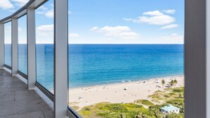Ein Balkon mit großen Fenstern und Blick auf einen Strand unter blauem Himmel.