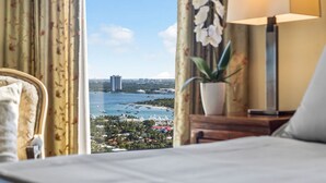 Ein Hotelzimmer mit Bett, Nachttisch mit Lampe, Blumentopf und Blick auf eine Küstenlandschaft mit Booten und Gebäuden.