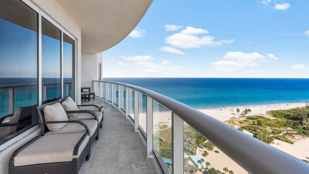 Ein Balkon mit Korbmöbeln und Glasgeländer mit Blick auf Strand und Meer.