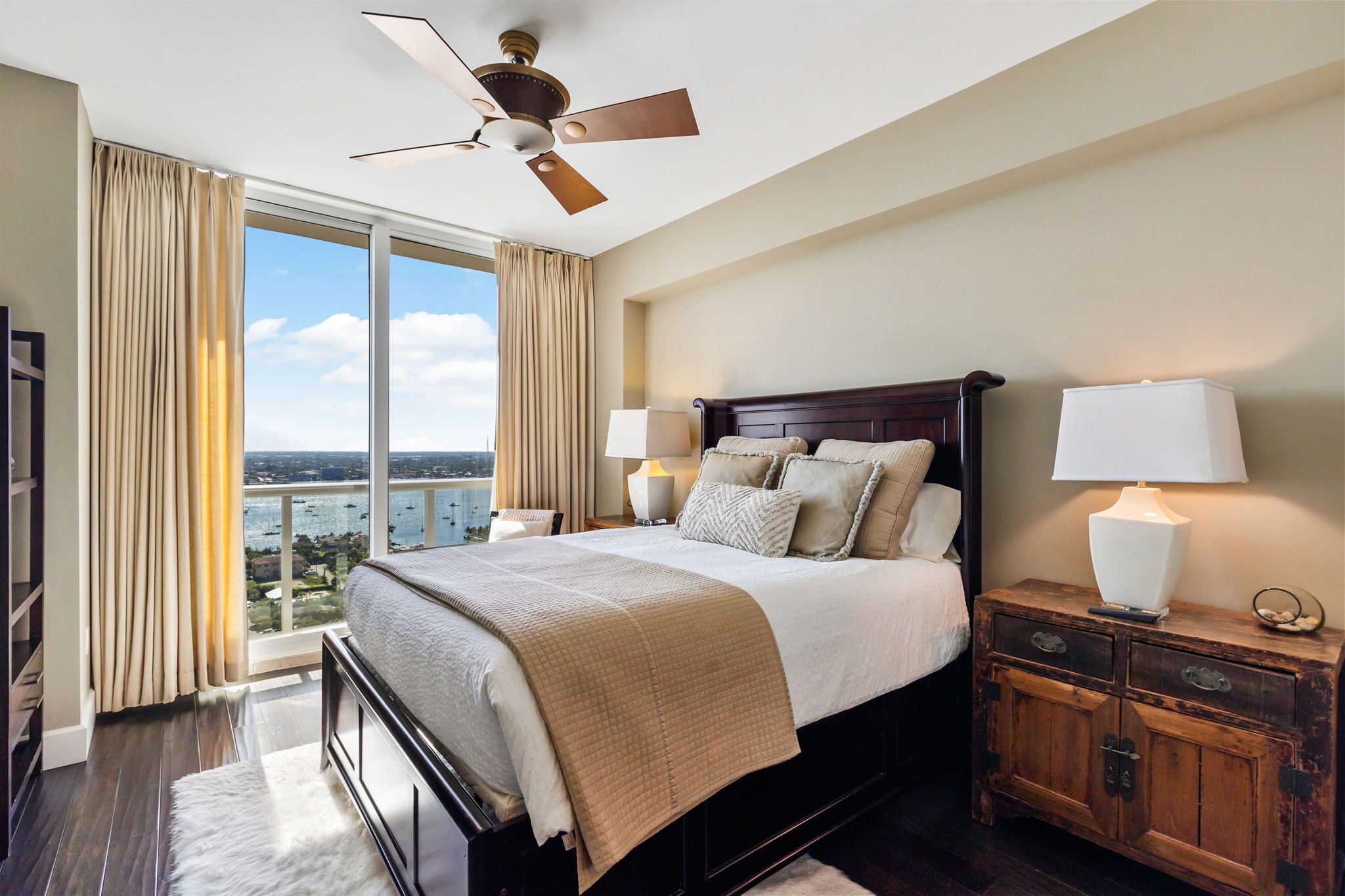 A bedroom with a large bed, two bedside lamps, a ceiling fan, and a view of the city through large windows.