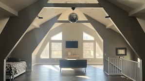 A spacious attic bedroom with a sloped ceiling, a ceiling fan, a large window, and a bed with a patterned bedspread.