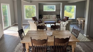 A spacious dining area with a large wooden table and chairs, a sofa, and a window with a view of greenery.