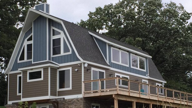 A two-story house with a prominent gable roof, a balcony with a wooden railing, and a brick foundation.
