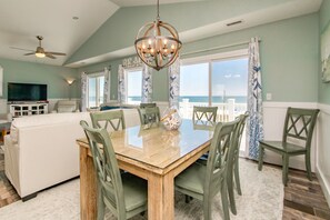 A dining area with a wooden table and chairs, a chandelier, and a view of the ocean through large windows.