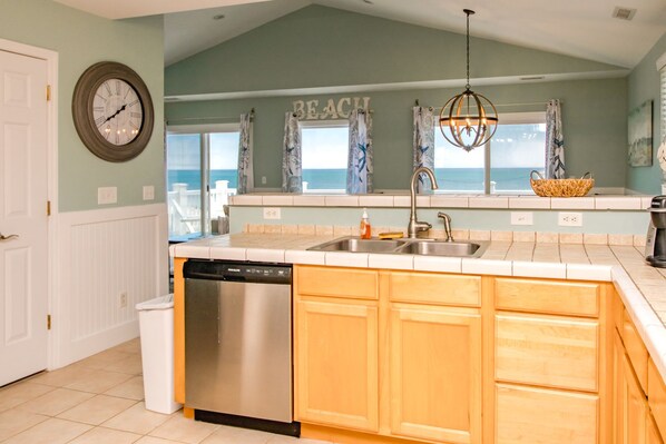A kitchen with a dishwasher, double sink, wooden cabinets, and a view of the beach through the window.