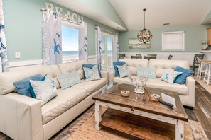 A living room with two beige sofas, a wooden coffee table, and a view of the beach through the window.