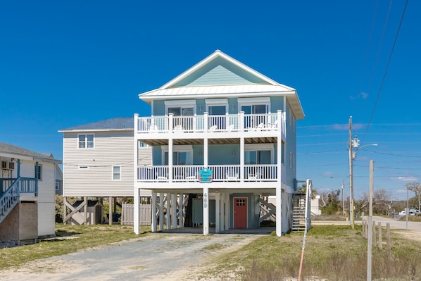 A two-story beachfront house with a balcony, a covered porch, and a red door.