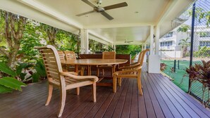 A covered outdoor patio with a round wooden table and chairs, surrounded by lush greenery and a view of a tennis court.