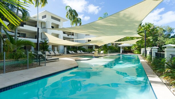 A swimming pool with lounge chairs and umbrellas, surrounded by palm trees and a building with balconies.