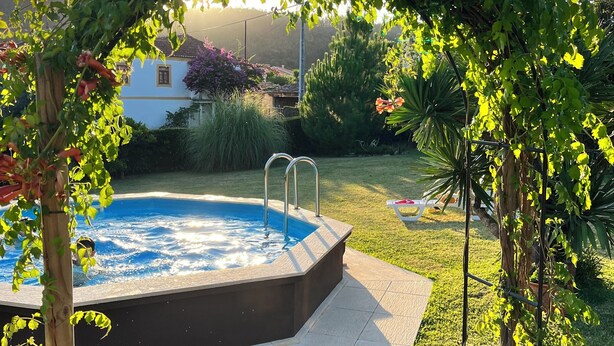 A swimming pool with a wooden deck, surrounded by lush greenery and a clear sky.