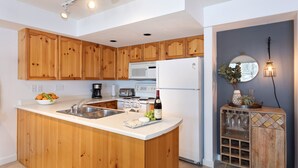 Una cucina con mobili in legno, un piano di lavoro bianco, un lavello incassato, un forno a microonde, un frigorifero e una bottiglia di vino.