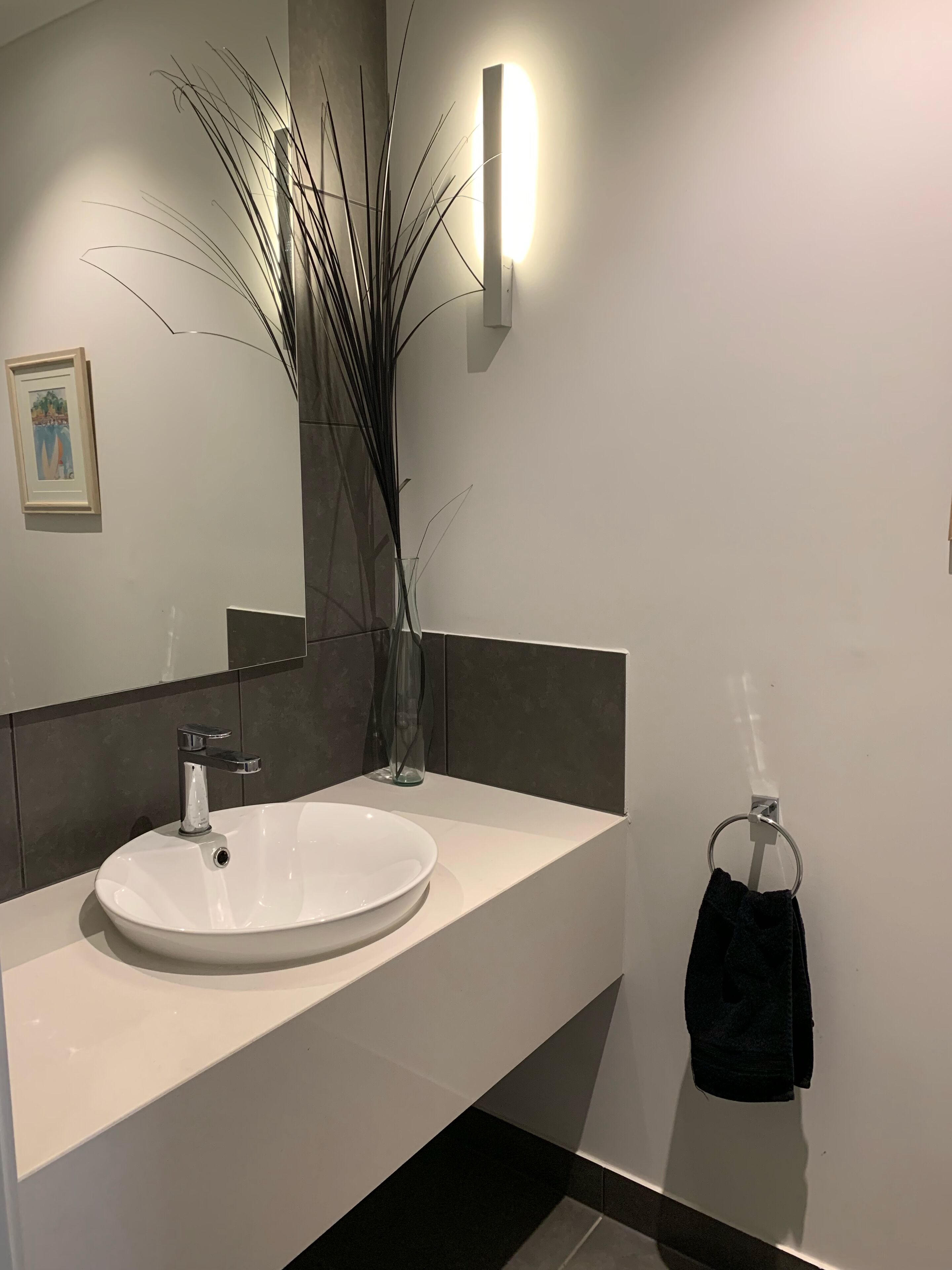 A modern bathroom with a white sink, a glass vase with tall plants, a towel ring, and a framed picture on the wall.
