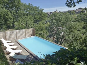 Una piscina rectangular con cubierta de madera y sillones de descanso, rodeada de abundante vegetación y con vistas a un pueblo histórico.