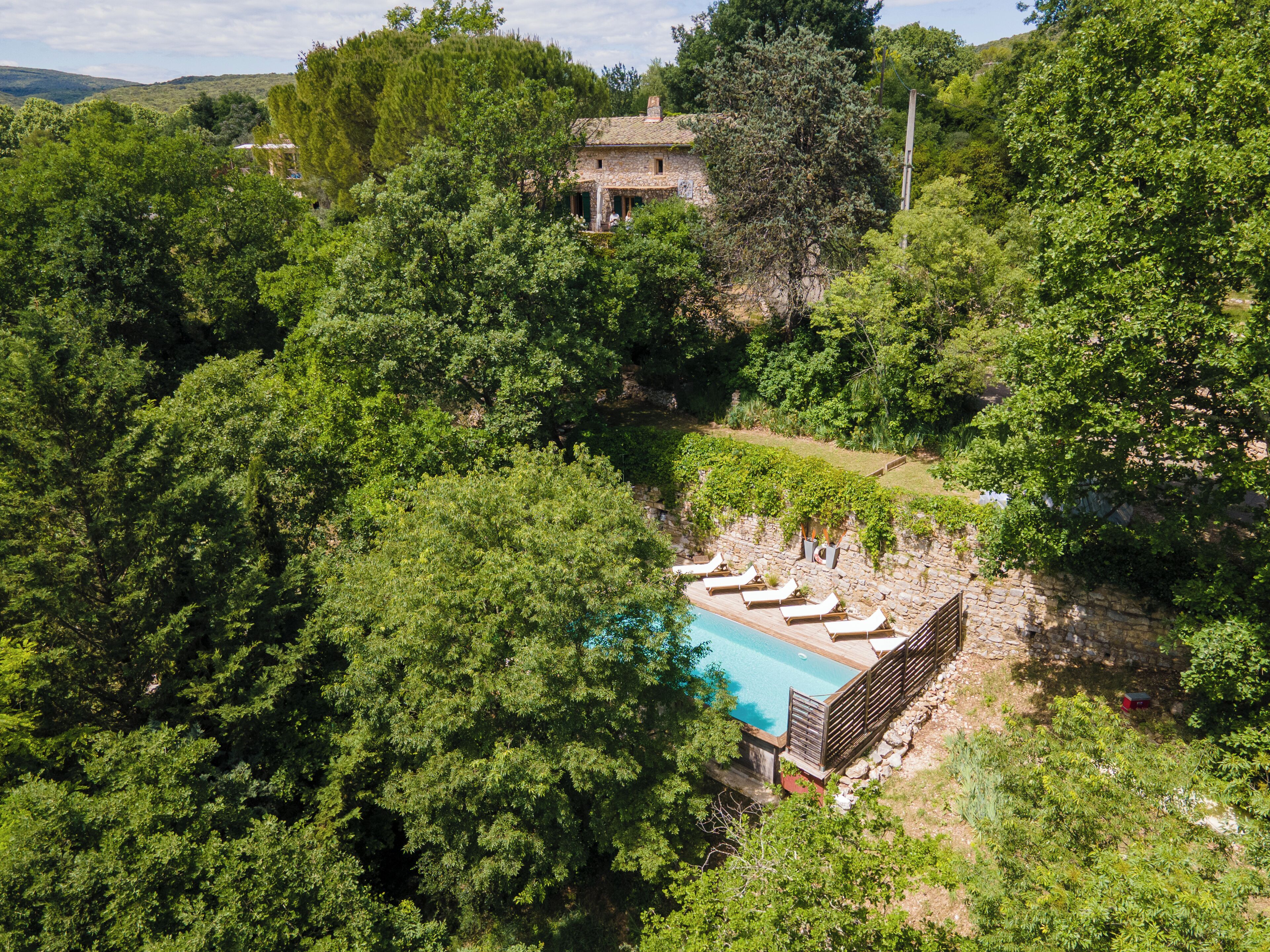A stone villa surrounded by dense forest, with a swimming pool and lounge chairs.