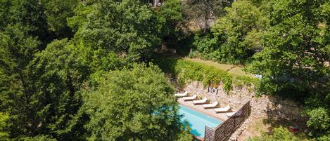 A stone villa surrounded by dense forest, with a swimming pool and lounge chairs.