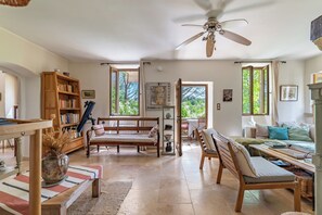 A spacious living area with a ceiling fan, a wooden bookshelf, a dining table with chairs, and a view through the open windows. - Stone farmhouse with pool (Saint-André-de-Roquepertuis)