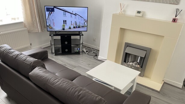 Uma sala de estar moderna com um sofá de couro marrom, uma mesa de centro branca, uma televisão sobre um móvel e uma lareira.