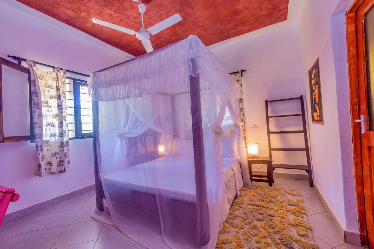 A bedroom with a four-poster bed, a ceiling fan, a bedside table with a lamp, a window with curtains, and a wooden door.