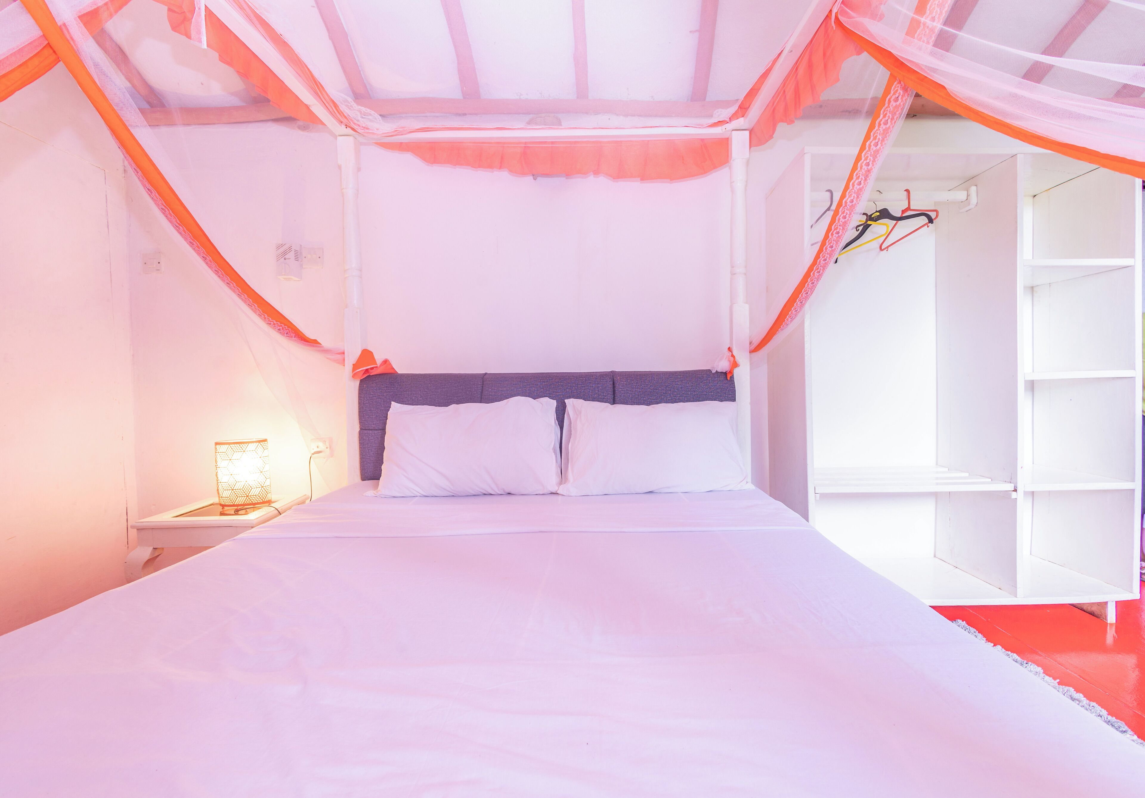 Ein Bett mit weißer Bettwäsche und einem orangefarbenen Moskitonetz darüber. Ein Nachttisch mit Lampe und ein Regal mit Kleidung sind ebenfalls vorhanden.