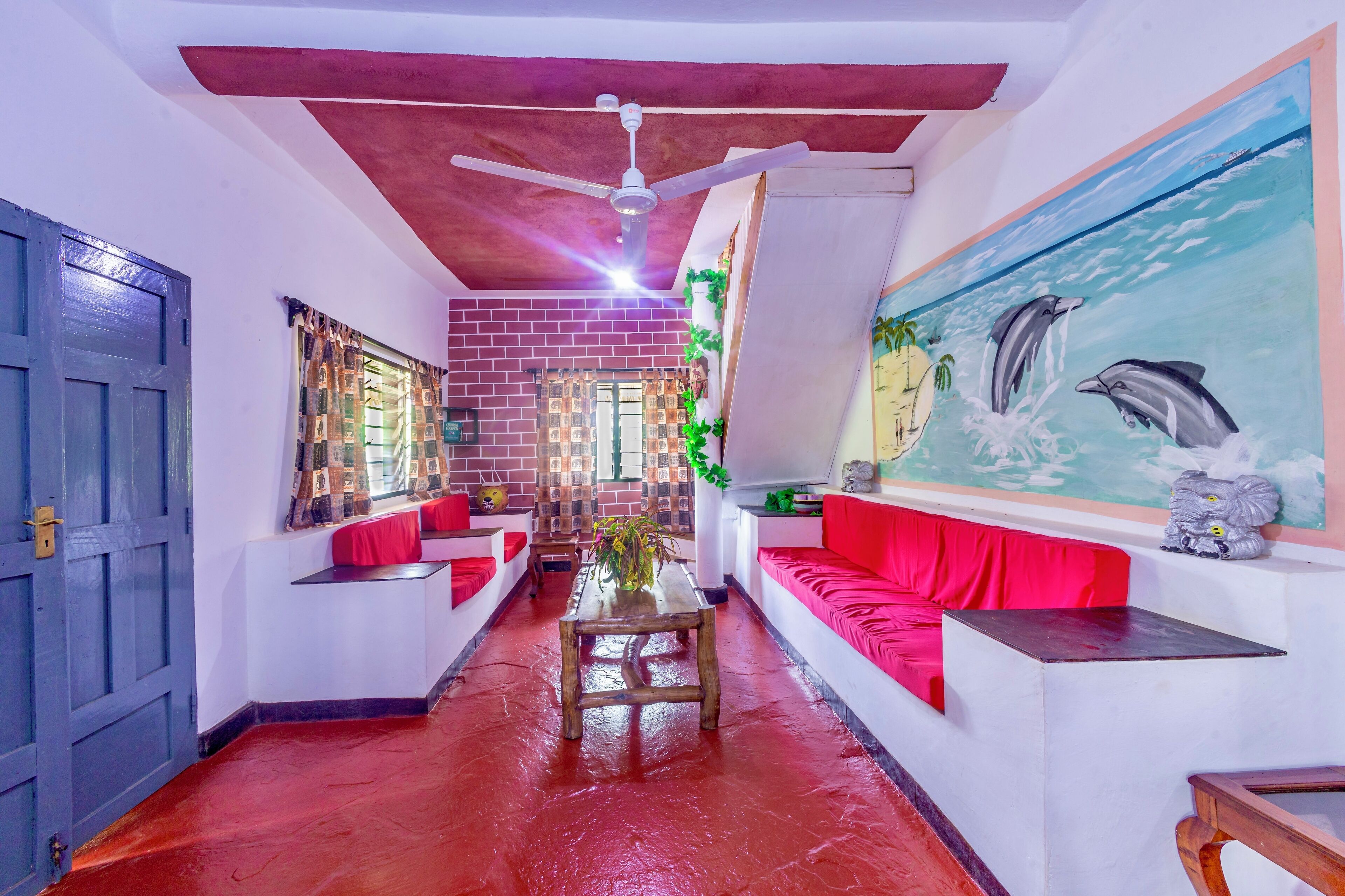 Un hall d’hôtel avec un sol rouge, un ventilateur de plafond, une grande fresque représentant des dauphins et un coin salon avec des coussins rouges.