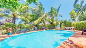 Una piscina rodeada de palmeras y vegetación, con una zona de descanso y un edificio pequeño al fondo.
