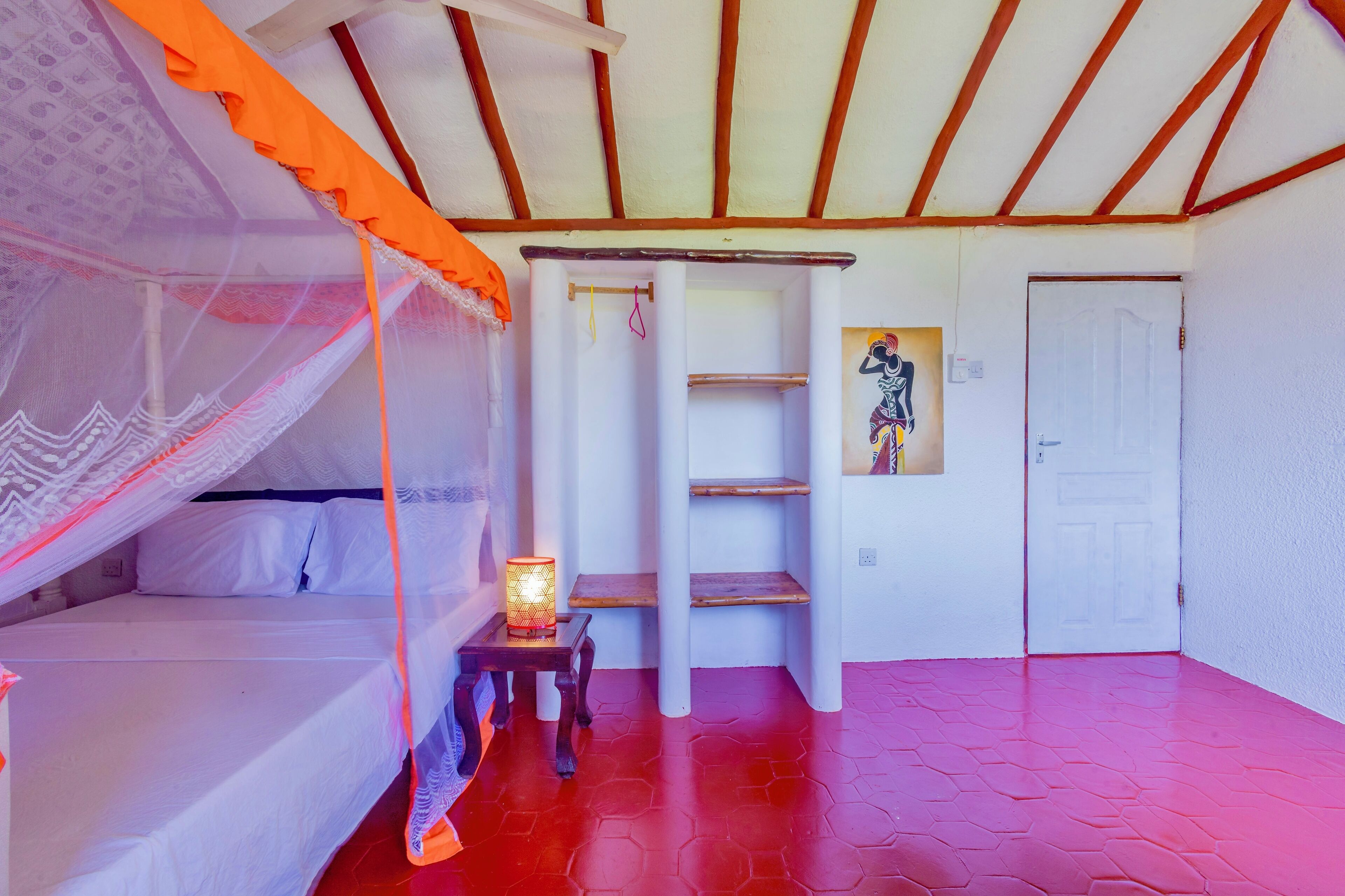 A room with a bed under a canopy, a small table with a lamp, a wooden shelf, and a door.