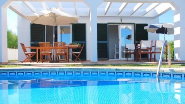 A swimming pool with a tiled deck, surrounded by wooden chairs and tables under a pergola.