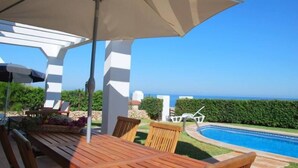 A wooden deck with a table and chairs, a large umbrella, and a pool with a view of the ocean.