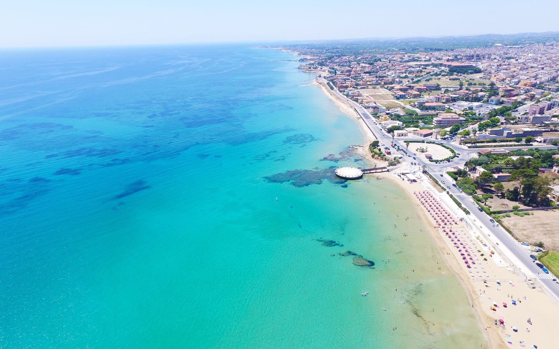 Vista aèria d'una ciutat costanera amb una platja, aigües blaves cristal·lines i una barreja d'edificis residencials i comercials.