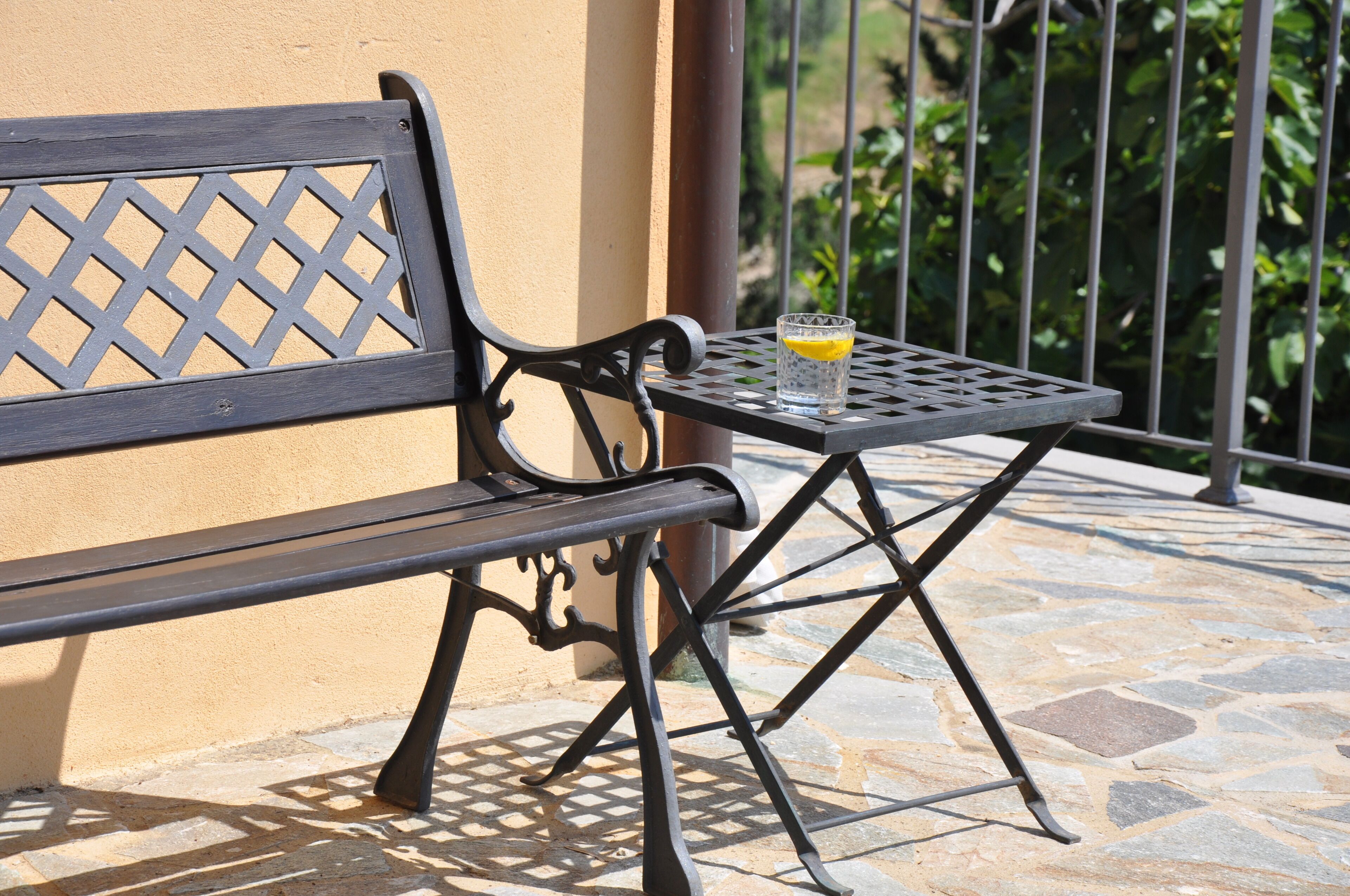 Un banc et une table en métal noir, avec un verre d’eau posé sur une terrasse carrelée en pierre.