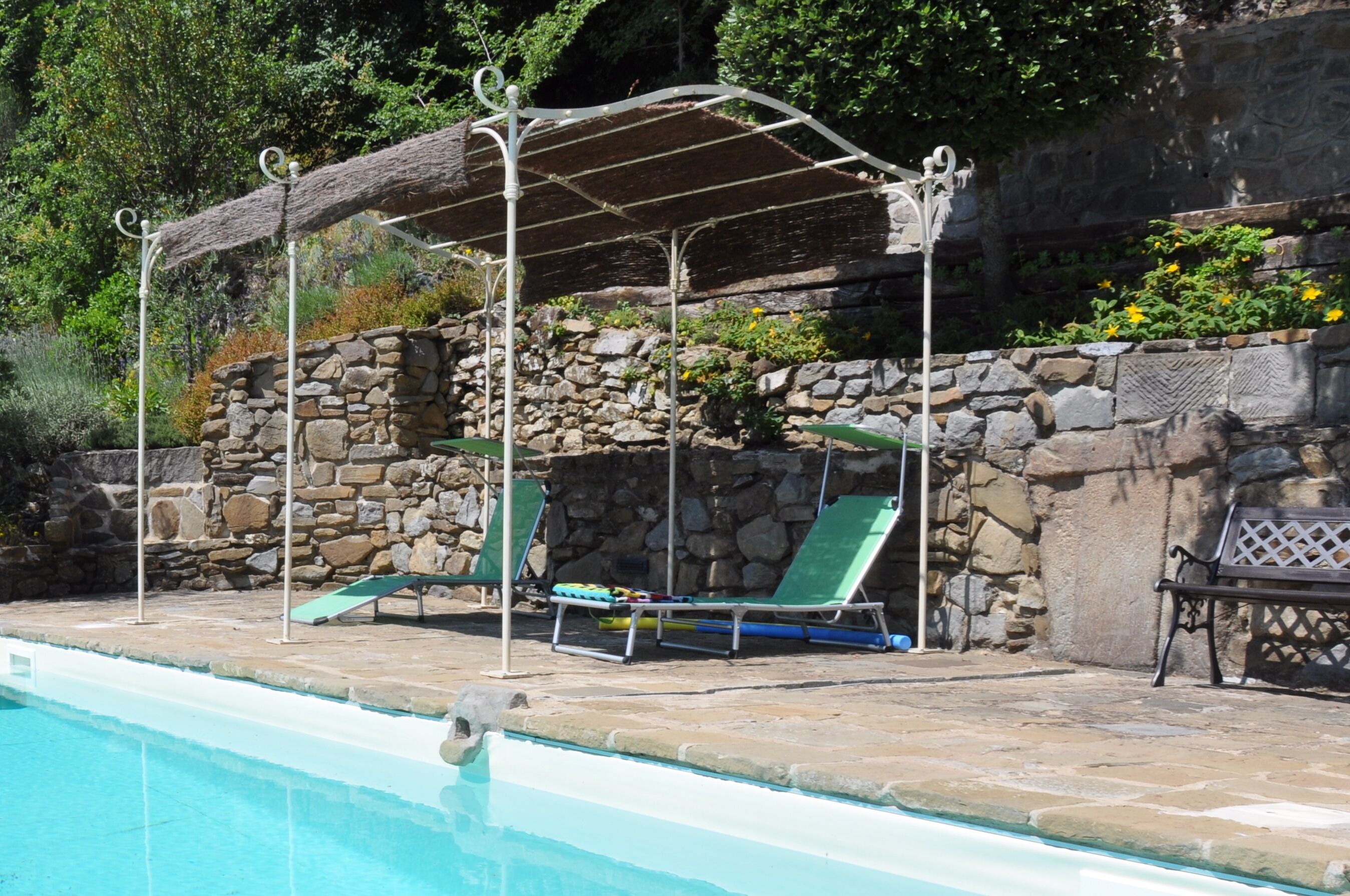 Un área junto a la piscina con un muro de piedra, un espacio de descanso cubierto y sillas de salón.