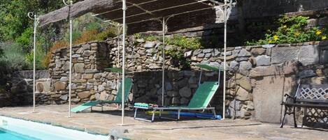 Un área junto a la piscina con un muro de piedra, un espacio de descanso cubierto y sillas de salón.