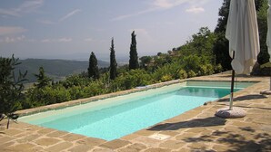 A rectangular swimming pool with a clear blue water, surrounded by stone tiles. There are tall trees and a view of distant hills in the background.