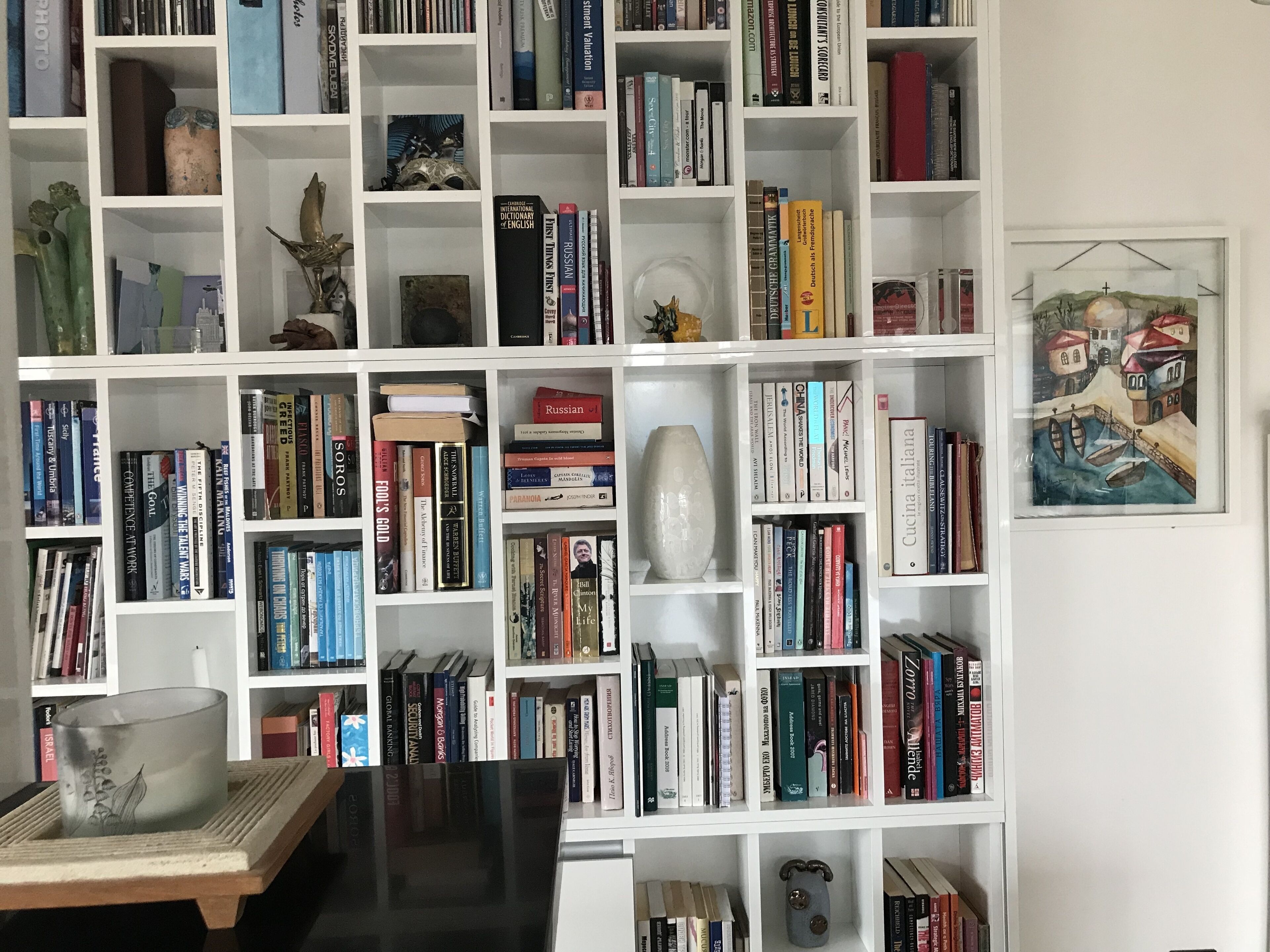 Une bibliothèque remplie de livres et d’objets décoratifs, un tableau encadré accroché au mur et une table sur laquelle se trouve un vase.